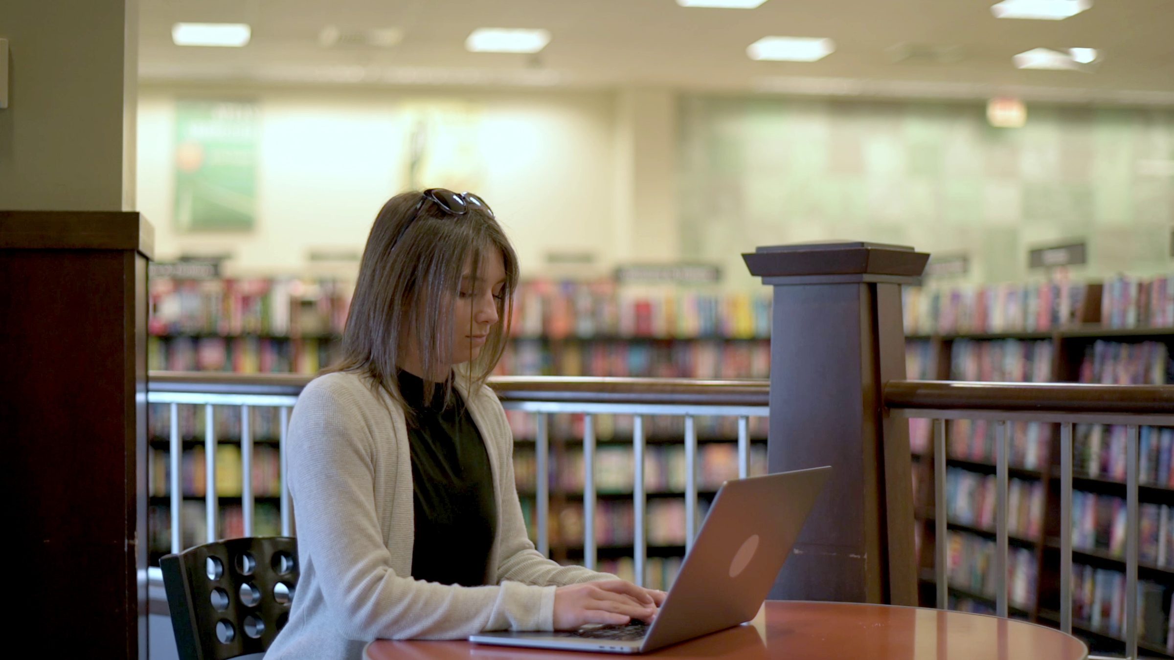A young woman types on a laptop at a round table, with library shelves blurred behind her