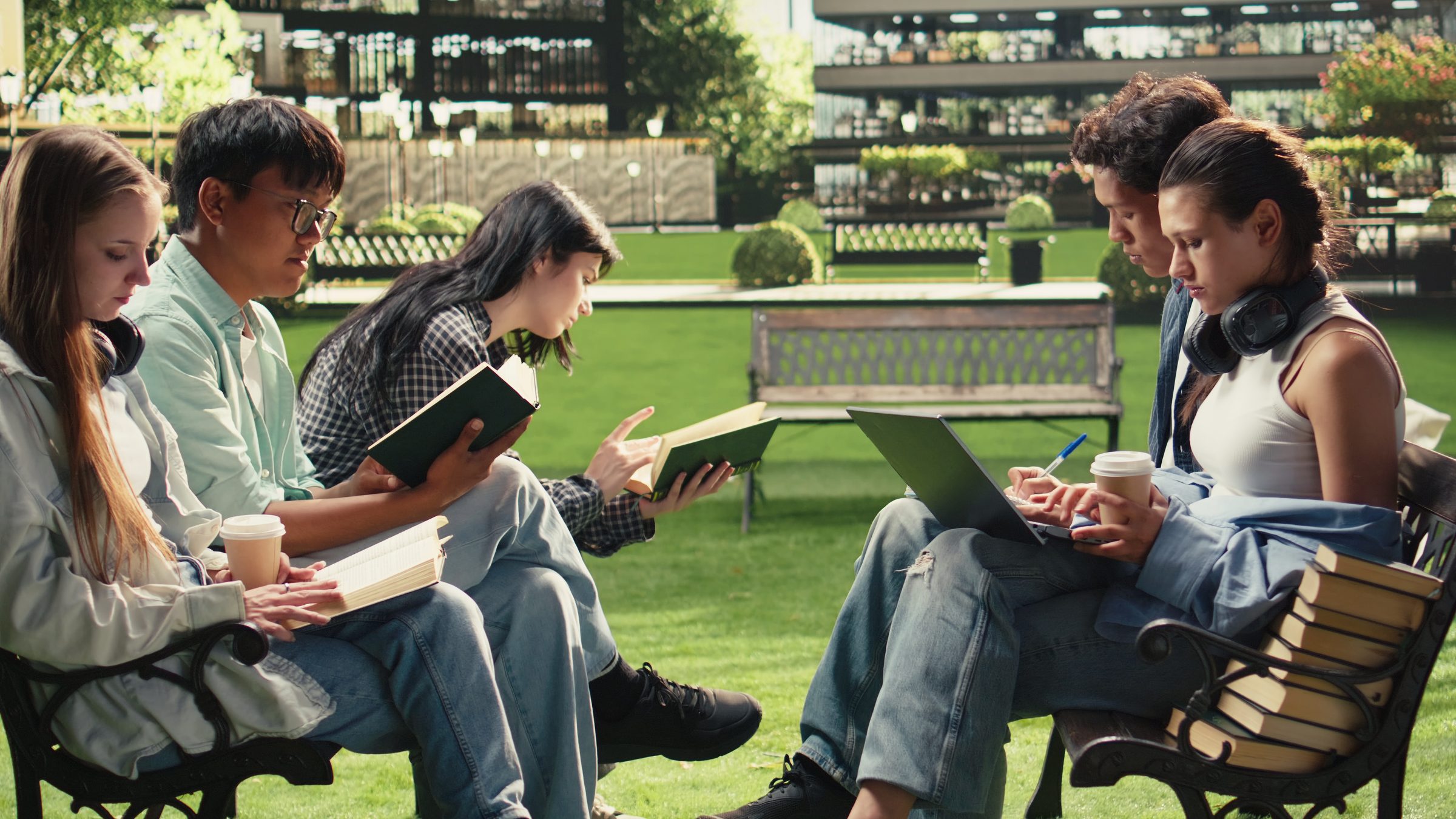 Five students sit on benches on a campus lawn with books, a laptop and coffee